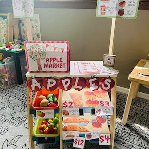 May include: A wooden play stand with a red and white checkered canopy. The stand has a sign that says "Apple Market" and a sign that says "Pay Here". There are shelves with toy apples, apple pie, candy apples, and apple butter. The price tags are visible, with prices ranging from $2 to $12.
