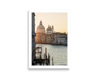 May include: A framed print of Venice, Italy, featuring a canal with wooden posts and buildings on either side. The image captures a sunset with warm, golden light illuminating the domes of a church in the background. The water reflects the sky.