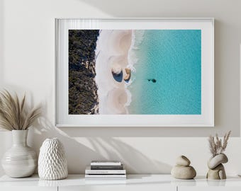 May include: Aerial photograph of a beach scene with white sand, turquoise water, and a strip of green trees. The image is framed in a white border and displayed on a white wall. Decorative vases and books are in the foreground.