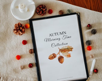 May include: A white printable planner with the text "AUTUMN Morning Time Collection" and a watercolour illustration of acorns and an oak leaf. The planner is surrounded by small brown and orange pom-poms and pine cones.