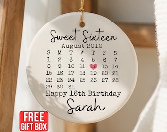 May include: A round white ceramic ornament featuring the text Sweet Sixteen, August 2010, and a calendar grid with a pink heart around the number 12. Below the calendar, the text reads Happy 16th Birthday and the name Sarah in black script.