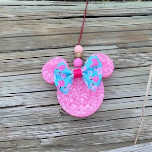 May include: Pink Minnie Mouse-shaped car air freshener with a blue bow decorated with pink flamingos. The air freshener is attached to a red string.