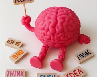 Könnte beinhalten: Eine leuchtend pinke, bewegliche Gehirnfigur mit Armen und Beinen, die ein kleines Holzschild mit der Aufschrift FEED ME hält. Um die Figur herum liegen sechs kleine rechteckige Holzplättchen mit dem Text USE ME, BRB, FIND ME, THINK, BUSY und IDEA.
