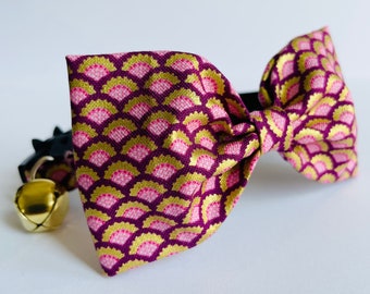 Puede incluir: Una pajarita de patrón rosa y dorado con una campana en un collar negro. La pajarita está hecha de tela con un patrón geométrico.