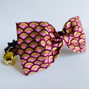 Può includere: Un papillon con motivo rosa e oro con un campanello su un collare nero. Il papillon è realizzato in tessuto con un motivo geometrico.
