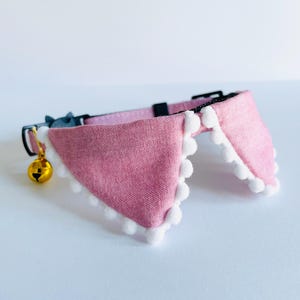 May include: A pink cat collar with a white pom-pom trim and a gold bell. The collar is made of fabric and has a black adjustable buckle.