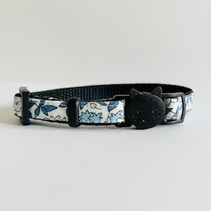 Puede incluir: Un collar para gato con estampado floral blanco y azul con una hebilla negra con forma de gato.