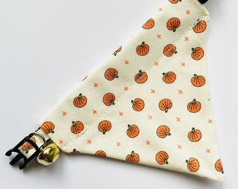 May include: A white fabric dog collar with a pumpkin pattern and a gold bell. The collar is adjustable and has a black buckle.