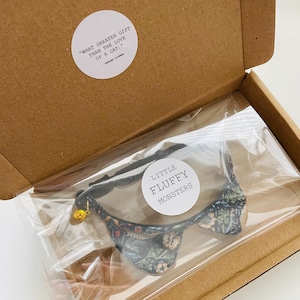 May include: A black cat collar with a floral pattern and a bell. The collar is packaged in a cardboard box with a quote about cats. The quote reads "What greater gift than the love of a cat." The brand name "Little Fluffy Monsters" is printed on a white circle sticker.