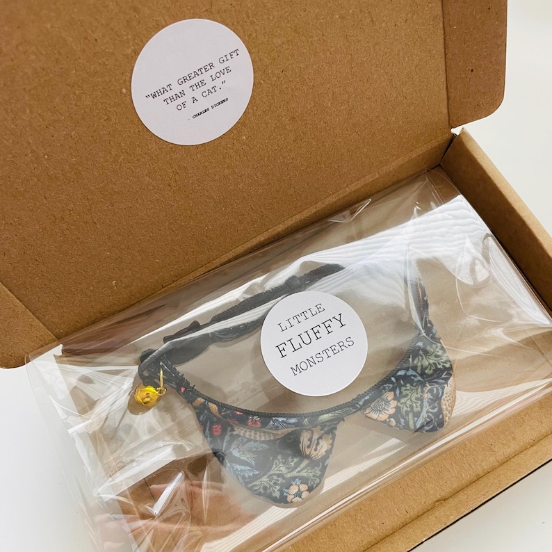 May include: A black cat collar with a floral pattern and a bell. The collar is packaged in a cardboard box with a quote about cats. The quote reads "What greater gift than the love of a cat." The brand name "Little Fluffy Monsters" is printed on a white circle sticker.