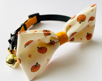 May include: A white fabric cat collar with a pumpkin print and a yellow bow. The collar has a gold bell attached.