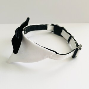May include: A white and black dog collar with a bow tie. The collar is made of a soft, white fabric with a black trim. The bow tie is made of black velvet.