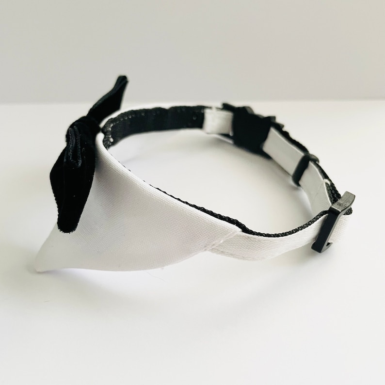 May include: A white and black dog collar with a bow tie. The collar is made of a soft, white fabric with a black trim. The bow tie is made of black velvet.