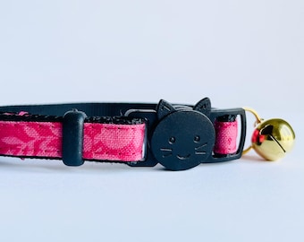 May include: A black cat collar with a pink floral pattern. The collar features a black cat-shaped buckle and a gold bell. The collar is designed for cats and is made of durable materials.