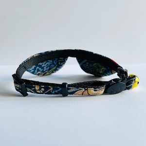 May include: A black cat collar with a floral pattern and a bell. The collar has a black buckle with a cat face on it.