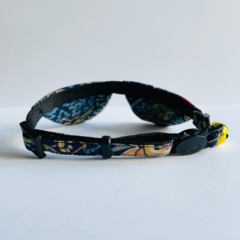 May include: A black cat collar with a floral pattern and a bell. The collar has a black buckle with a cat face on it.