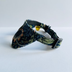 May include: A black cat collar with a floral pattern in shades of green, blue, and pink. The collar has a black buckle and a small bell.
