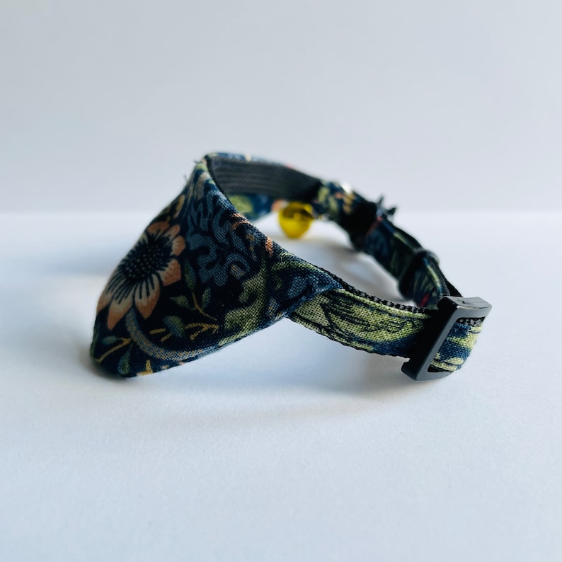 May include: A black cat collar with a floral pattern in shades of green, blue, and pink. The collar has a black buckle and a small bell.