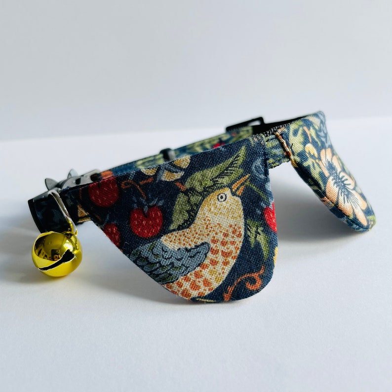 May include: A blue cat collar with a floral pattern featuring birds and berries. The collar has a bell attached.