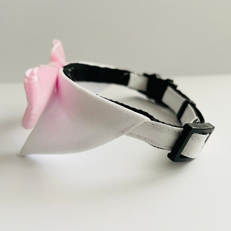 May include: A white and pink dog collar with a bow tie. The collar is made of fabric and has a black adjustable buckle.