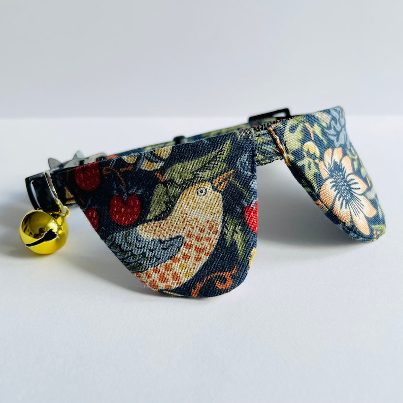 May include: A blue cat collar with a bell and a floral pattern featuring birds and berries. The collar has two large, triangular flaps.