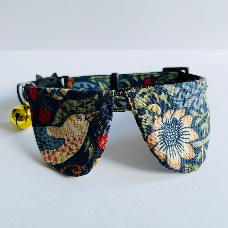 May include: A blue and green floral patterned cat collar with a bell and two large, pointed, fabric flaps. The flaps are decorated with a bird and a flower.