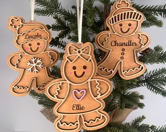 May include: Three wooden gingerbread person ornaments with personalized names. The ornaments are brown with white and pink details. The names are "Lincoln", "Chandler", and "Ellie".