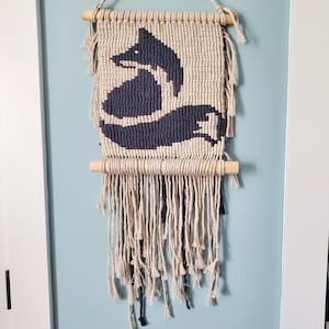 May include: A black and white macrame wall hanging featuring a fox design. The hanging has a wooden dowel at the top and a fringe of yarn at the bottom.