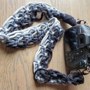 May include: A camera strap made of a blue and black crocheted material with a metal ring. The strap is attached to a black camera with the word "Canon" printed on it.