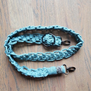 May include: A teal blue crocheted camera strap with a black metal ring and two black metal clasps. The strap has a brown leather patch with a turtle design.