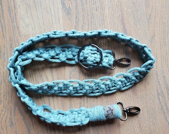 May include: A teal blue crocheted camera strap with a black metal ring and two black metal clasps. The strap has a brown leather patch with a turtle design.