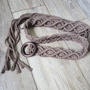 May include: A brown macrame belt with a wooden ring buckle. The belt is made of thick, braided cord and has a simple, bohemian style.