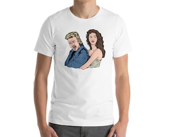 Può includere: Una maglietta bianca con un'immagine di un uomo e una donna in stile cartone animato. L'uomo indossa una giacca blu e ha i capelli biondi. La donna ha i capelli lunghi e castani e indossa un vestito bianco.