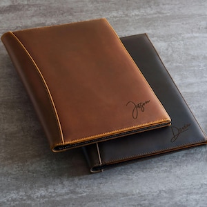 May include: Two leather portfolios, one in medium brown and one in dark brown, featuring stitched edges. The medium brown portfolio is engraved with the name Jason, and the dark brown portfolio is engraved with the name Devin in script font.