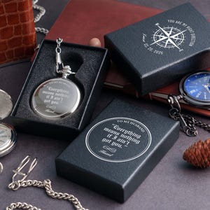 a collection of items, including a silver pocket watch, a black box, a blue watch, and a pine cone, all placed on a dark surface.