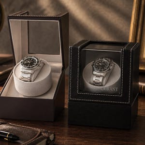May include: Two black watch display cases with glass lids, each containing a silver-toned wristwatch with a black dial on a light gray cushion. The right case features white stitching and the text L.GRANT printed on the glass front panel.