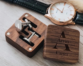 Puede incluir: Un par de gemelos redondos de tono plateado con la letra A grabada, dentro de una caja cuadrada de madera oscura con tapa magnética. La tapa tiene grabado el nombre Arvin Carter y una letra A grande. Un reloj de pulsera con correa negra.