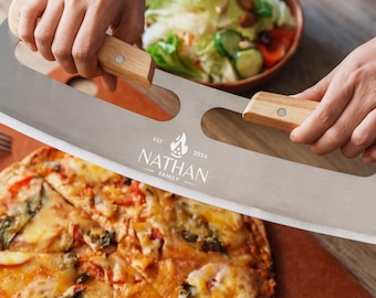 May include: A stainless steel pizza cutter with two light wood handles. The blade features an engraved logo with a flame icon, the text EST 2024, and the name NATHAN FAMILY. The tool is positioned over a sliced pizza on a brown surface.