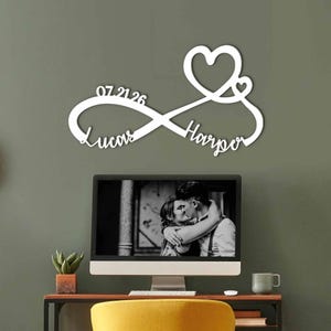Op de afbeelding: Een wit wandbord in de vorm van een oneindigheidssymbool met een hart aan de bovenkant, de namen Lucas en Harper in cursief schrift en de datum 07.21.26 geïntegreerd in het ontwerp als decoratief item voor aan de muur.