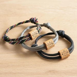 May include: Three adjustable cord bracelets featuring wooden beads engraved with crossed lines, letters, symbols, and numbers. One bracelet has a multicolored cord, one is grey, and one is black, each with a sliding knot closure mechanism.
