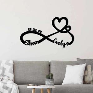 May include: A black wall-mounted sign featuring an infinity symbol shape. The design incorporates a large heart, a smaller heart, the date 12.24.25, and the names Ethan and Evelyn integrated into the continuous flowing lines of the metal structure.