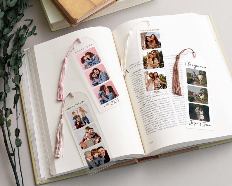 Four customized bookmarks with different family images, messages, and colors resting on an open book with a eucalptus vine off to the side of the desk.