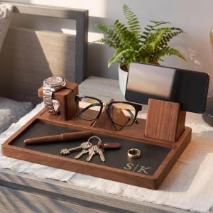 May include: A dark wood desk organizer featuring a rectangular tray with a black inlay, engraved with the letters S|K. It includes a vertical stand for a smartphone, a raised block for a wristwatch, a central pyramid-shaped holder for eyeglasses, a pen, a set of keys, and a ring.