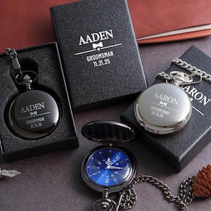 a collection of three black pocket watches, each with a personalized engraving, placed on a dark surface alongside a pen and a pine cone.
