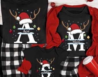 May include: A family of four wearing matching black and white plaid pajamas with Christmas reindeer designs. The shirts feature the names "Ace", "Mom", "Jeremy", and "Sofia" with a Santa hat on each reindeer.