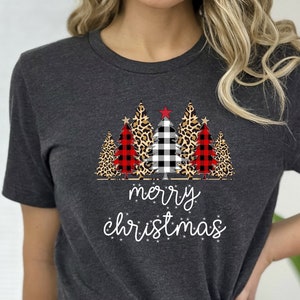 May include: A dark grey t-shirt featuring a Christmas tree design. The trees are decorated with red and black buffalo check, black and white gingham, and leopard print. The text "merry christmas" is printed below the trees.