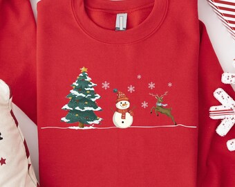 May include: A red crewneck sweatshirt featuring a Christmas tree, snowman, and reindeer design. The festive graphic is embroidered in green, white, and brown. The sweatshirt is surrounded by Christmas decorations, including a pillow with the words "CLAUS NORTH POLE".