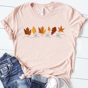 May include: A pink t-shirt featuring a line of autumn leaves in orange, yellow, and brown shades. The leaves are connected by a thin black line.