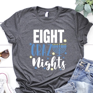 May include: A dark gray t-shirt with the text "Eight Crazy Nights" in white and blue. The text is surrounded by small yellow stars. A menorah with blue and yellow flames is in the center of the text.