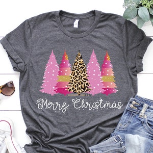 May include: A charcoal grey t-shirt with a Christmas tree design. The trees are pink, gold, and leopard print, with the words "Merry Christmas" in white script. The shirt is styled with blue jeans and white trainers, creating a casual festive look.